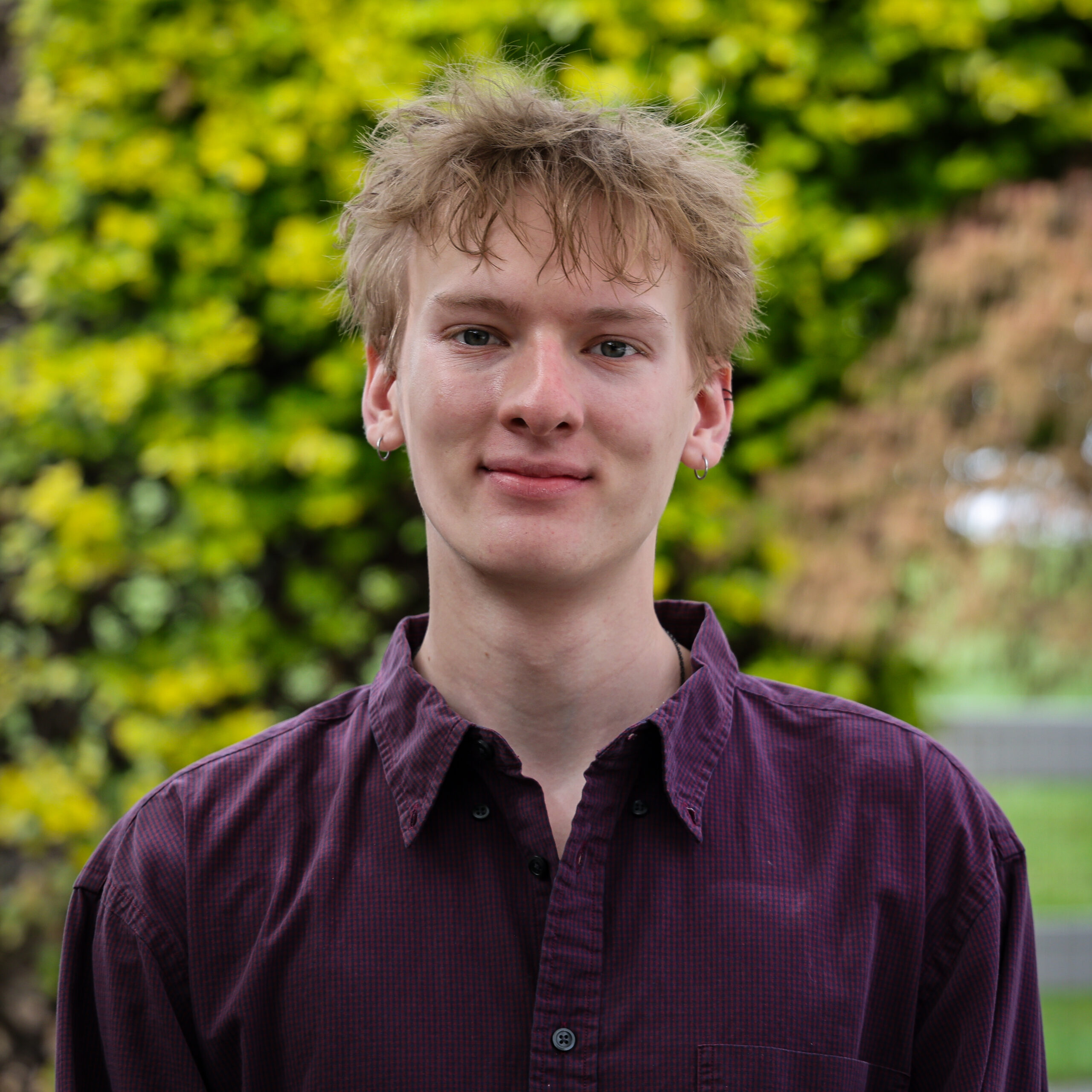 A portrait photograph of a young man with blonde hair, wearing a dark purple pinstripe shirt, with silver hoop earrings, against a background of green hedges