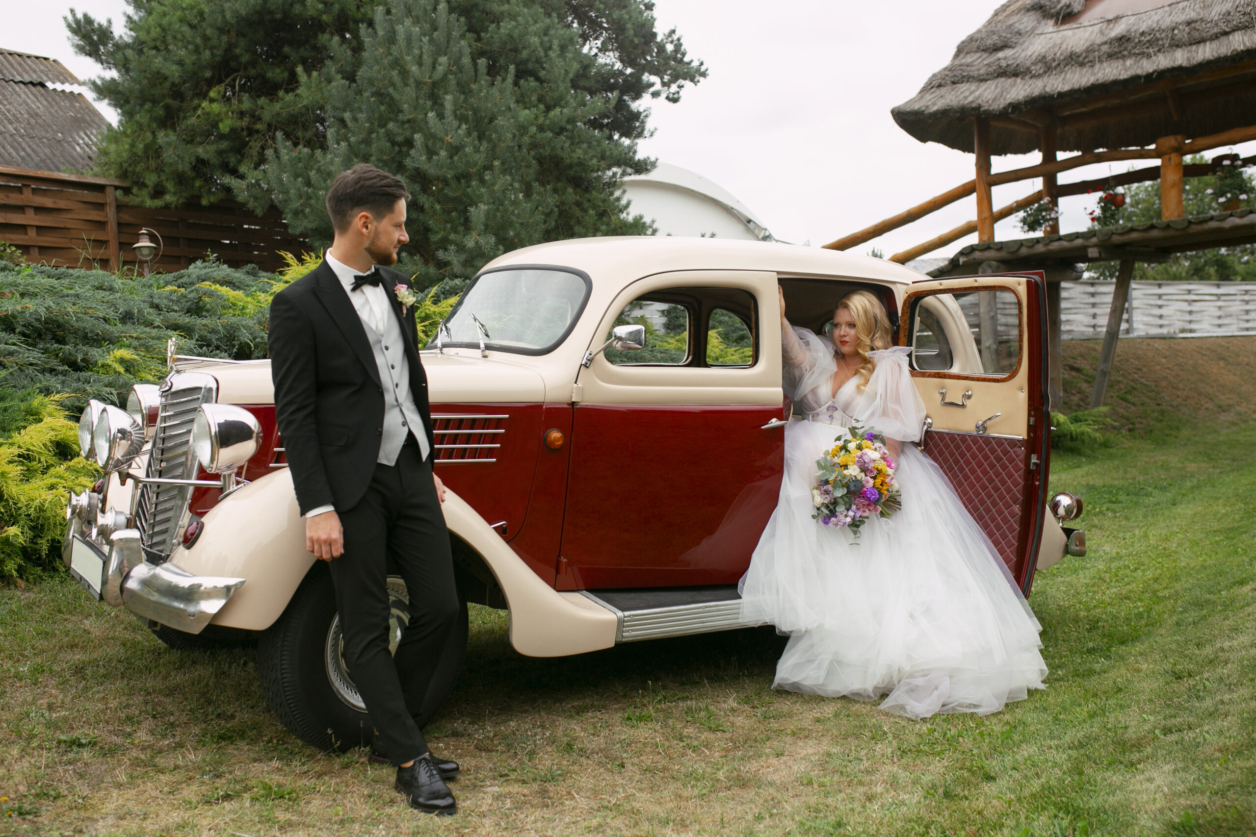 Groom in a tuxedo looking at a vintage wedding car at an outdoor venue